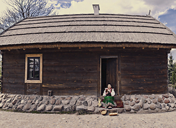 drewniany budynek przed którym siedzi dziewczyna, trzymająca kurę