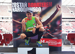 mężczyzna w stroju sportowym kuca na pierwszym stopniu podium 34. warszawskiego maratonu na piersi ma zawieszony medal