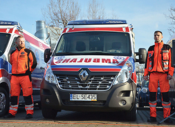 przód ambulansu po obu stronach stoją dwaj ratownicy medyczni