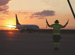 Samolot kołujący po płycie lotniska o zmierzchu. Na pierwszym planie sterujący ruchem z lampami w uniesionych rękach