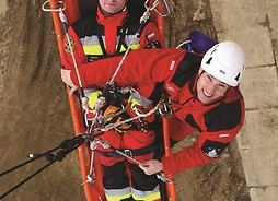 Dwóch ratowników na ćwiczeniach – jeden leży na noszach zawieszonych na linie, obok drugi