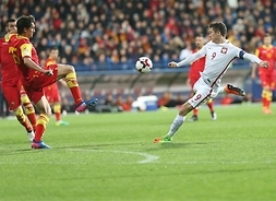 Piłkarz w stroju reprezentacji Polski z opaską kapitańską kopie piłkę z powietrza prawą nogą. Przed nim próbujący blokować strzał rywal