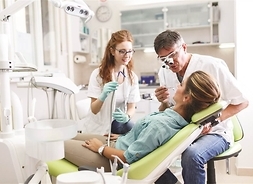 Kobieta siedzi na fotelu dentystycznym. Nad nią, z przyrządami stomatologicznymi pochylają się kobieta i mężczyzna w fartuchach.
