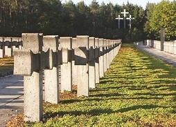 Widok na cmentarz, z rzędem murowanych identycznych krzyży. W tle pod lasem trzy duże krzyże