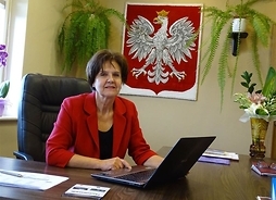 Samorządowiec przy biurku i laptopie, w garsonce, z tyłu duże godło Polski