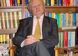 Zdjęcie starszego mężczyzny w garniturze i krawacie siedzącego na fotelu przed biblioteczką