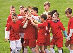 Grupa dzieci w piłkarskich strojach wznoszących do góry puchar. Stoją na murawie boiska