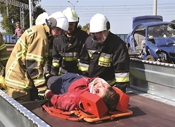 Trzech strażaków w bojowym umundurowaniu układa na noszach rannego