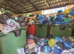 Wiata śmietnikowa z przepełnionymi kontenerami na śmieci. Worki ze śmieciami rozsypanymi przed kontenerami