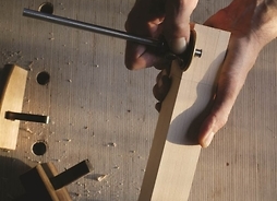Na zdjęciu widoczne są dłonie stolarza, który trzyma kawałek drewna oraz narzędzie stolarskie