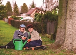 Mężczyzna i kobieta siedzą na trawie. Przed nimi stoi konewka i leżą grabki. Mężczyzna trzyma jabłko w ręku.
