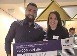 Mężczyzna i kobieta trzymają przed sobą symboliczny czek - nagrodę w konkursie Mazovian Startup