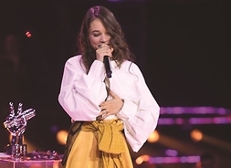 Młoda dziewczyna stoi na scenie, trzyma w dłoni mikrofon