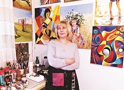 kobieta w fartuchu stoi w swojej pracowni na ścianie, za jej lecami  wiszą obrazy