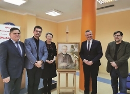 Elegancko ubrani mężczyźni i jedna kobieta stoją obok obrazu Jacka Malczewskiego. Obraz umieszczony jest na sztalugach.