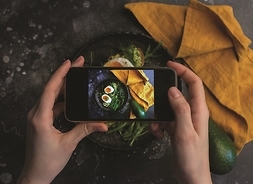 widok na dłonie trzymające smartfon, za pośrednictwem którego robione jest zdjęcie potrawy