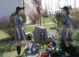 Dwie dziewczynki w mundurach harcerskich stoją na baczność przy kamieniu pamiątkowym. Na kamieniu umieszczono tabliczkę z informacją historyczną. Przy kamieniu mała sadzonka dębu.