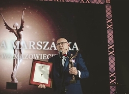 Ryszard Nowaczewski na scenie, prezentuje otrzymaną nagrodę Marszałka Województwa Mazowieckiego, wraz z dyplomem.