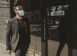 Mężczyzna w masce ochronnej, w okularach przeciwsłonecznych i w garniturze przechodzi obok zamkniętego lokalu