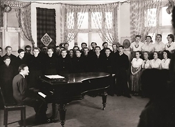 W dużym planie - Tadeusz Sygietyński prowadzi zajęcia chóru „Mazowsza” w sali prób - czarno-białe zdjęcie.