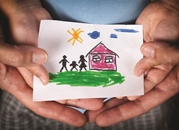 Dłonie dziecka i osoby dorosłej trzymające rysunek przedstawiający dom i rodzinę