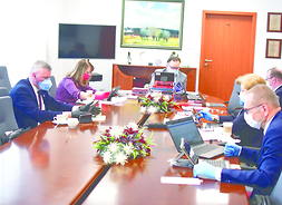Członkowie Zarządu Województwa Mazowieckiego oraz skarbnik województwa siedzą po dwóch stronach długiego stołu i uczestniczą w obradach  posługując się laptopami i tabletami.