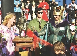 Dwie uśmiechnięte kobiety w królewskich koronach i kostiumach z epoki, pośrodku którch stoi figurka rycerza.