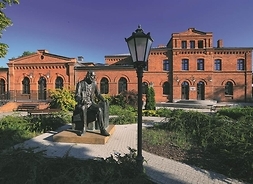 Budynek z czerwonej cegły, przed którym znajduje się pomnik mężczyzny, w postawie siedzącej