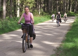 Kobieta jadąca na rowerze leśną ścieżką rowerową. W tle grupa rowerzystów.