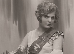 Młoda kobieta w eleganckiej jasnej sukni siedzi na fotelu. Włosy ma ułożone w fale, nie patrzy w obiektyw. Z lewego ramienia na prawe biodro spływa jej szarfa z herbem Radomia i napisem 1932. Na kolanach ma złożony bukiet kwiatów.
