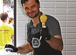 Uśmiechnięty mężczyzna w fartuchu trzymający w ręku rożek z lodami gałkowanymi.