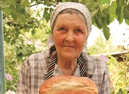 Kobieta w chustce na głowie trzymająca w obu dłoniach bochenek chleba.