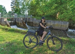 Dziewczyna na rowerze, pozująca do zdjęcia. Zdjęcie w plenerze, na tle tamy i płynącej rzeki.
