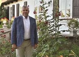 elegancki mężczyzna pozuje do zdjęcia na tle chałupy w skansenie, stoi przy kwiatach