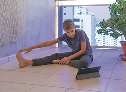 chłopiec siedzi na podłodze (siad płotkarski) próbuje dłonią sięgnąć palców stóp patrzy w ekran tabletu, który stoi przed nimi patz