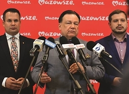 przed mikrofonami stoją trzej mężczyźni w garniturach, w tle ścinka z logo Mazowsza