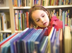 Dziewczynka stoi przy półce z książkami i ogląda grzbiety książek