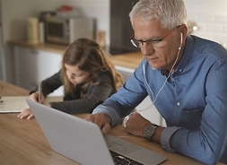 Przy biurki siedzi starszy mężczyzna, w uszach ma słuchawki. Patrzy w ekran usatwionego przed nim laptopa. Obok siedzi mała dziewczynka i pisze coś w zeszycie.