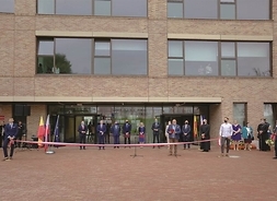 budynek szkoły, przed którą stoją elegancko ubrani ludzi, przy nich mikrofony