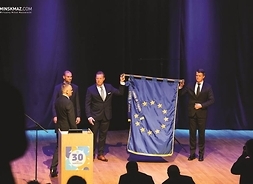 na scenie stoi czterech mężczyzn w garniturach, dwóch z nich trzyma flagę UE