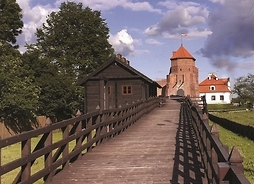 Zdjęcie zrbione z pozycji osoby stojącej na drewnianym moście. W oddali, na drugim końcu mostu jest wieża zamkowa.