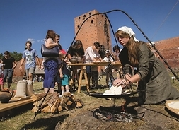 Młoda kobieta w średniowiecznym stroju przygotowuje nad paleniskiem podpłomyki