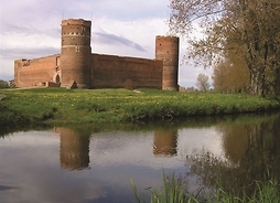 Zdjęcie zamku z czerwonej cegły, z dwiema wieżami. Obok nurt rzeki.
