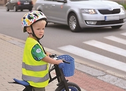 chłopiec na rowerze w kamizelce odblaskowej i kasku stoi przed pasami, przed którymi widać samochód osobowy