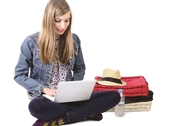 na podłodze siedzi dziewczyna, na kolanach ma laptop, obok koce i kapelusz