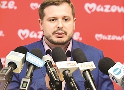 mężczyzna z lekkim zarostem stoi na tle ścianki z logotypami Mazowsze mówi do mikrofonów różnych stacji radiowych i telewizyjnych