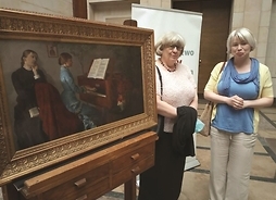 Obraz jacka Malczewskiego w grubej ramie stoi na sztalugach. Obok niego stoją dwie kobiety.