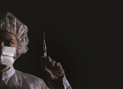 Pielęgniarka w stroju zabiegowym z maseczką chirurgiczną na twarzy trzyma uniesioną lewą rękę. W dłoni trzyma strzykawkę zwróconą igłą do góry