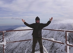mężczyzna z podniesionymi rękoma pozuje do zdjęcia na szczycie góry, w tle krajobraz górski