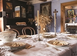 Wnętrze chaty z XIX w. N pierwszym planie stoi stłó przyjryty jasnym obrudem N stoie dwie świece, rozstawiona zasatawa stołowa